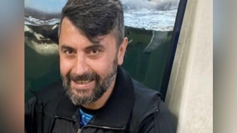 Cumali Turhan has black hair swept to one side and a greying, black beard. He is wearing a black jacket and appears to be sitting in front of an aquarium display