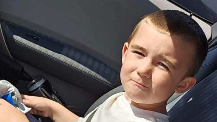 Lincoln Button is sitting in a car. He is squinting into the sun as he poses for the camera. He has cropped brown hair and is wearing a white T-shirt.