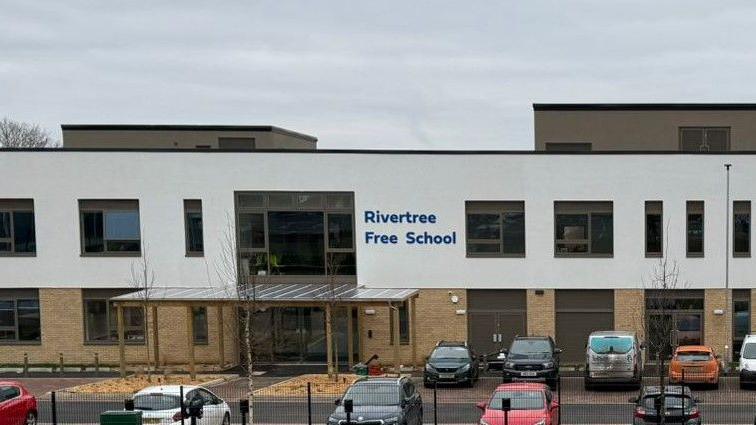 The front of a school which has two storeys and cars parked outside. 