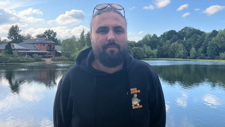 James Duffy stood in front of the lido. He has dark hair and a dark beard. He is wearing a black hoodie with BIG DUFFS FISHING on the front.