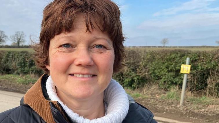 Louise James, looking straight at the camera, smiling, wearing a white jumper and black coat, with a brown trim around the neck. She has short dark hair and is standing by a road that has a bush behind her, with a small sign. 