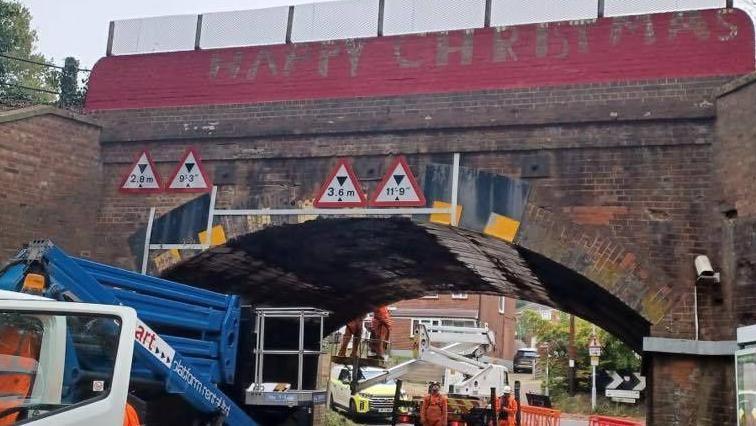 The Christmas Bridge being repainted by people in high vis clothing. They are standing on a platform being raised by a piece of machinery.