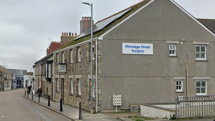 A stone building on a street with a white sign, and the words Meneage Street surgery in blue lettering