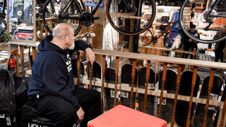 A man with a grey beard sits, leaning on the wooden balustrade of a mezzanine level, surrounded by skateboards and bicycles