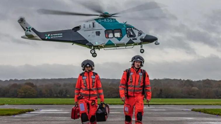 Two crew members from the Air Ambulance Charity Kent Surrey Sussex wearing orange flying kit with black helmets on a runway with a blue and white helicopter very low behind them.