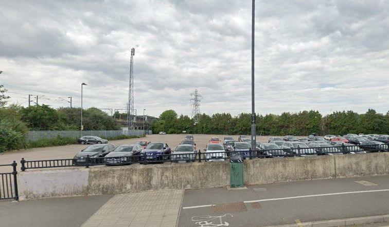 The car park seen from the road, with about 40 cars parked in it in daylight. There are trees lining one edge and a metal fence along the left border, and the overhead wires and gantries of the train line.