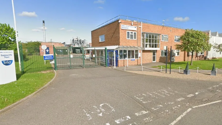 The exterior of the Tetley factory. It is a brick building behind a green gate. A Tetley sign can be seen on the road in front.