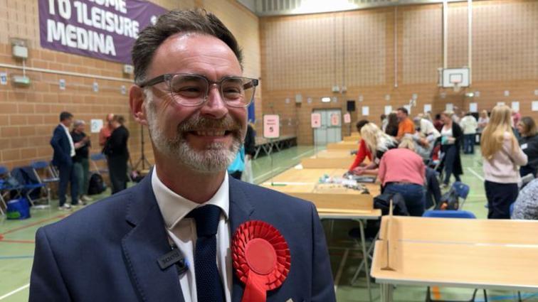 Richard Quigley smiles looking away from the camera, he has short hair and wears large square grey framed glasses with a short full beard. He wears a dark blue suit, with a white shirt and dark blue tie and red Labour rosette pin. People can be seen counting votes in the background of a sports hall.