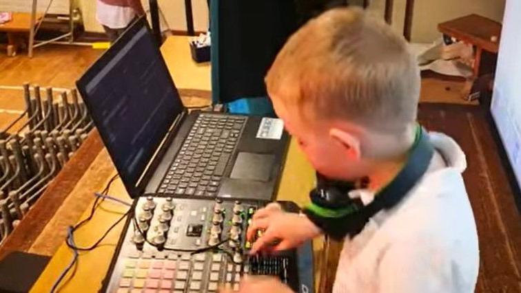 A younger Fionn O'Kane is pictured wearing a white polo shirt and a pair of black of headphones around his neck. He is mixing and is staring down at a laptop and a controller