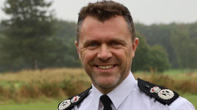 A man with brown hair is in a white shirt with black badges on the shoulders and a black tie.