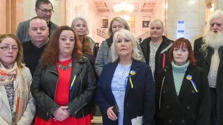 A group of 10 people stand facing a camera. There are three men and seven women. Behind them is a large white marble hall. Some of them wear blue and yellow pins