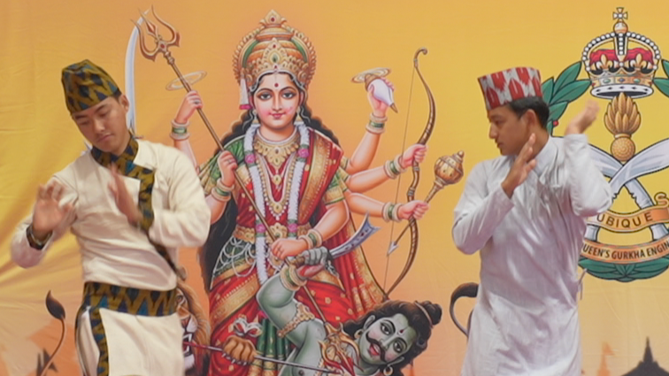 Two men wearing traditional Nepalese suits and hats dance on a stage. Behind them is a background featuring several Hindu gods.