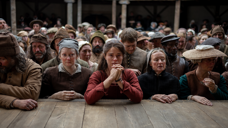 A film still from Hamnet, showing Jessie Buckley as Agnes (Anne Hathaway). The picture shows a group of characters who are audience members, pressed against the stage of the Globe. Agnes is in the middle in a red dress with her hands pressed together, looking somber.