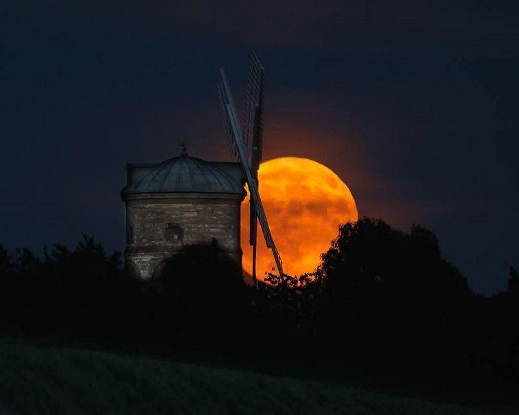 A bright orange, red Moon is illuminated behind the sails of a windmill with trees outlined against the sky.