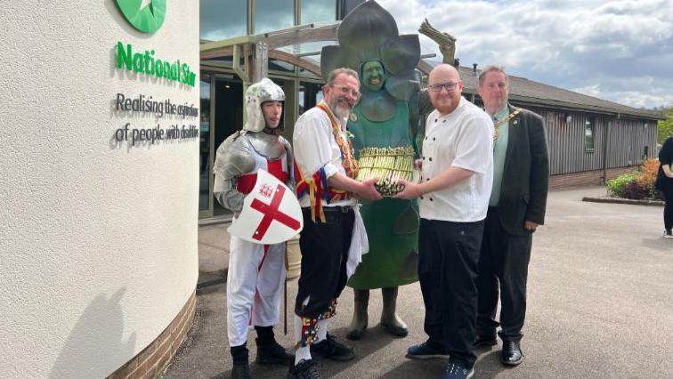 The giant asparagus standing in the middle of a group of people holding a bundle of asparagus