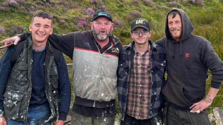 Jacob Kelly, Paddy O’Neill, Jake Mayers and Nic Tinnion stand side-by-side smiling and with their arms around each other. They are wearing outdoor clothing that is covered with white paint stains.