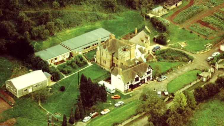 An aerial picture of Berrow Wood School with fields surrounding a black and white Tudor manor. There are cars parked on the road outside and bushes and trees around the edge