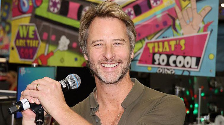 Chesney Hawkes smiling and holding a microphone in a studio. He has short fair hair and a beard. He is wearing a brown shirt and a poster behind him says You're So Cool in graffiti style writing.