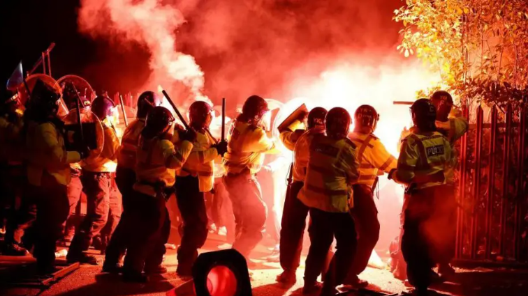 Officers in riot gear holding up batons and circular shields as red flares are thrown at them