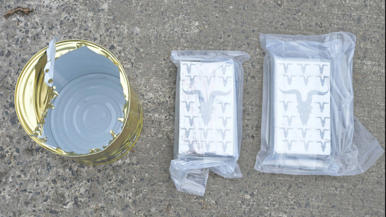 An opened, empty gold food can sits on the left while two rectangles of suspected drugs are wrapped in plastic on the right. All three items are placed on the concrete ground.