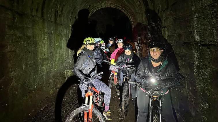 A group of woman in bikes wear helmets and warm jackets as they pose for a night-time picture in a tunnel. One of the cyclists is holding a torch.