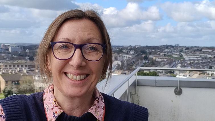 Rachel Goodsell taking a selfie on a rooftop. She has short brown hair and purple glasses and is wearing a pink floral shirt and a navy tank top. 