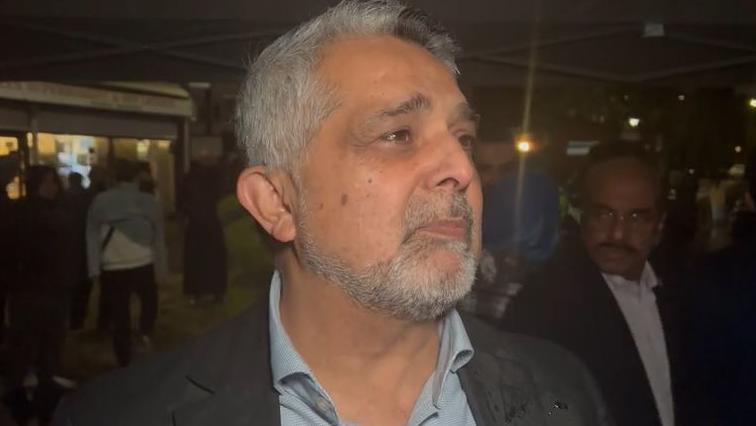 A middle aged man with grey hair and beard speaks to a reporter. It is night time and there are people in the background.