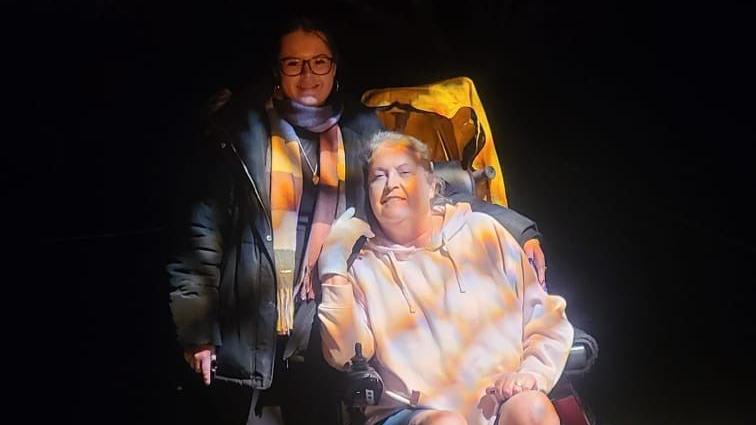 Jordan Till and her mum at a light exhibition. The background is dark and there is a light pattern signing onto the two people. Jordan's mum is in an electric wheelchair and Jordan is stood to her right with her arm around her. 