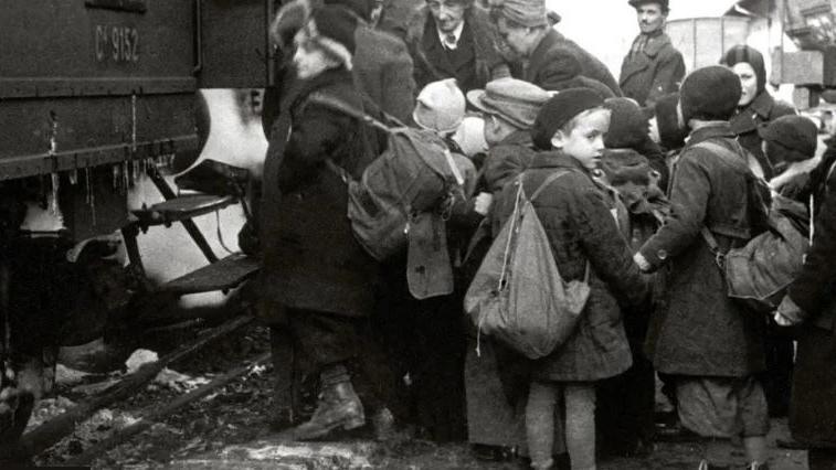 A black and white image of children boarding a train in 1940s. There are about a dozen children, with a handful of adults in the background. 