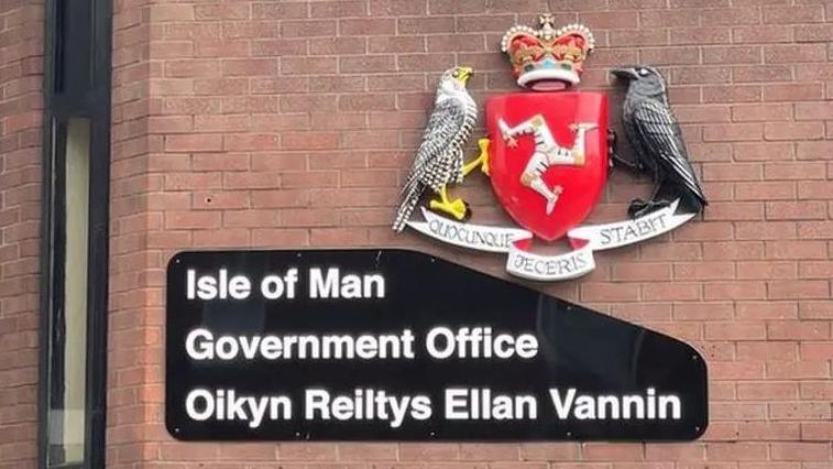 A sign on a red brick wall that reads Isle of Man Government Office in English and Manx on a black background next to the government crest.