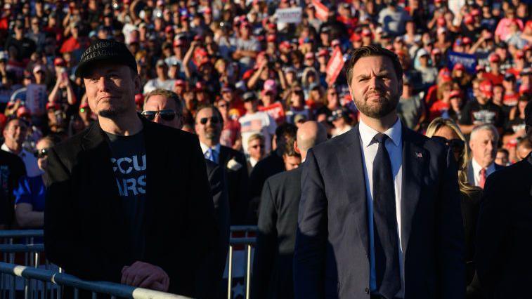 Elon Musk and JD Vance look ahead, in front of a crowd, with sun on their faces. 