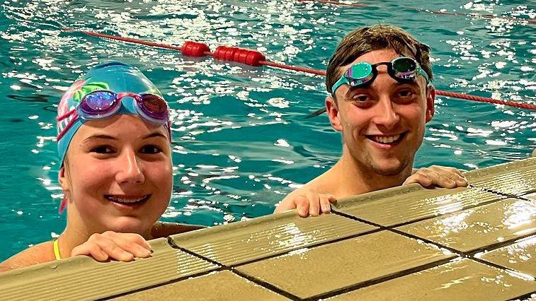 Swimmers Pemberley and Harvey are in the pool with just their heads and hands out of the water. Pemberley is wearing a blue swimming hat with her goggles on top. Harvey has his goggles pushed up on his brown hair. 