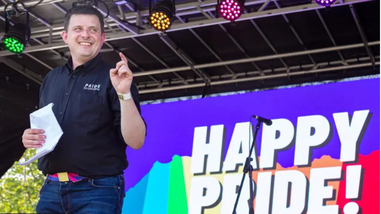 A man in a blue shirt and jeans is standing on a stage with a sign that reads "Happy Pride" in the background.