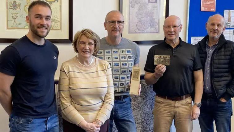 A young man in his 20s wears a blue t-shirt and jeans, standing next to one elderly woman and three older men at a library. Two men in the middle are holding WW1 postcards