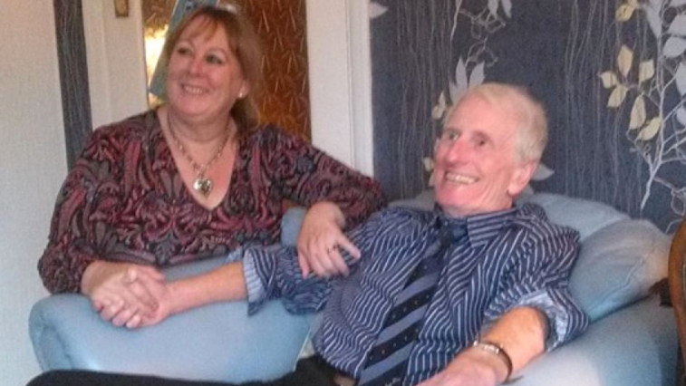 Dorothy and Melvin Cook. Melvin has short, light grey hair. He is wearing a blue stripe shirt and tie. He is sitting in an armchair. Dorothy is sat next to him, holding his hand and his arm. Both of them are smiling and looking over to the side. 