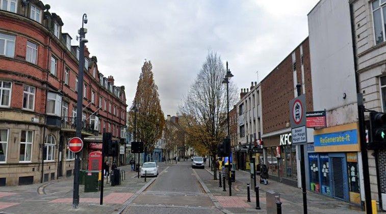 A town centre high street with traffic lights, trees, shop fronts and other street furniture. There are some parked cars but no pedestrians about.