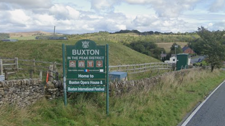 Green sign which reads "Buxton in the Peak District" in front of wooden fencing and green fields