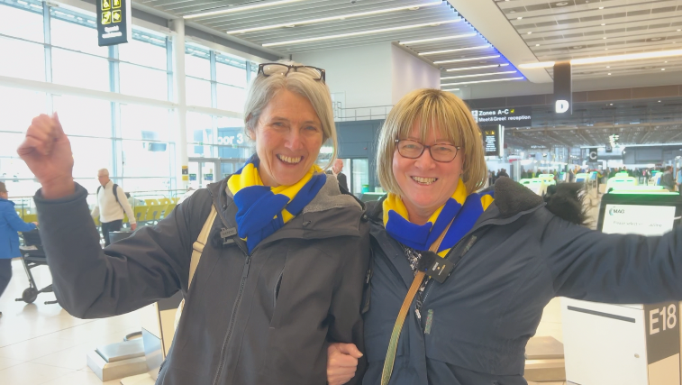 Carolyn (left) has her arm linked with Jeanette's. They are both wearing dark jackets and blue and yellow striped scarves. Carolyn has grey hair pulled back from her face and black glasses on her head. Jeanette has blonde bobbed hair, a fringe and wears dark-rimmed glasses.