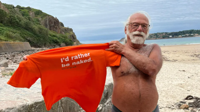 A topless Ross Mantle, with white hair, white beard and glasses, on a beach, holding up a T-shirt saying "I'd rather be naked"