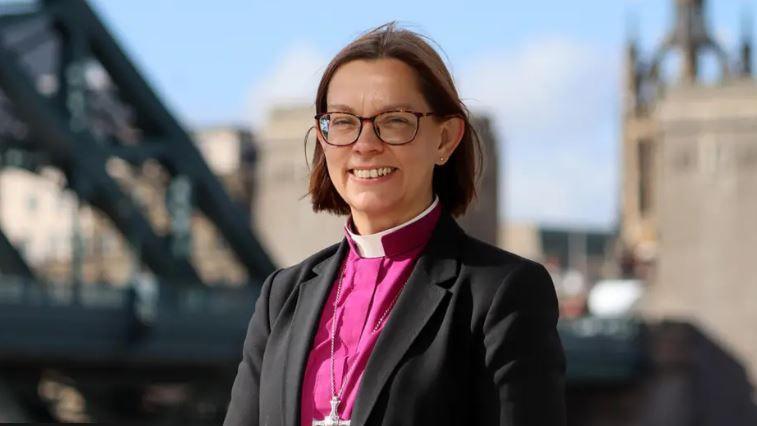 Helen-Ann Hartley wears a clerical collar with a pink shirt, black blazer, and silver cross chain, in a picture taken on the streets of Newcastle