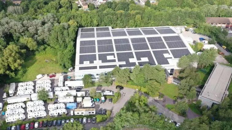 An aerial view of Bottle Yard studios showing a large warehouse roof covered in solar panels and surrounded by trees, there appears to be seven rows of trailers in the bottom left corner of the frame.