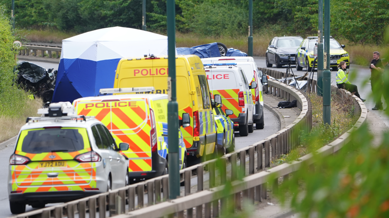 Police vans of varying size are lined up in a row leading up to the sie of the wreckage, all with reflective liveries. To one side, two police officers rest with paper cups, with one sat on the metal barrier between the two sides of the road.