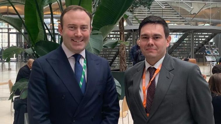 Blake Stephenson is in a navy double-breasted suit, wearing a tie and pink shirt. Beside him is a man with a grey suit and spotted tie. They are standing in Portcullis House at Westminster.