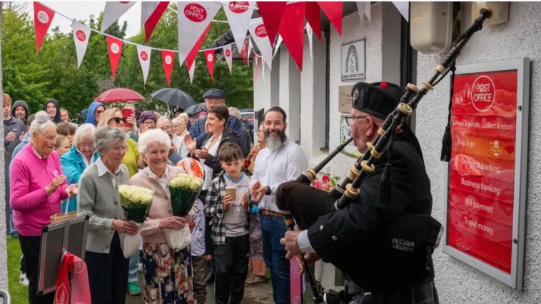 Cuirm ann an Cille Chòmhain airson ath-fhosgladh Oifis a' Phuist. Tha daoine cruinn còmhla le flùraichean agus tha pìobaire ann. 