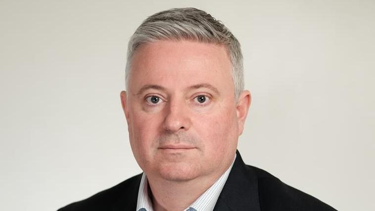 Paul Scott looking at the camera with a serious expression. He is wearing a black suit jacket and a white shirt without a tie. He has short grey hair.