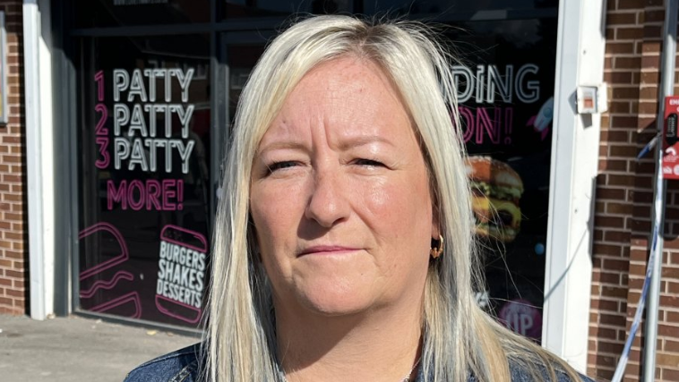 Louise is standing outside the takeaway. She has long blonde hair and a denim jacket on. The shop has black advertising signs filling the windows on either sides of the entrance. It is promoting burgers.