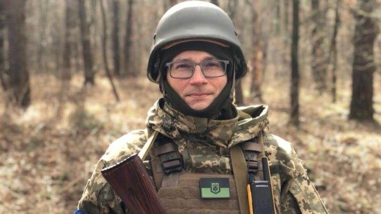 A man dressed in combat gear stands in a wooded area. He looks directly at the camera. He is wearing an army hard hat and glasses. You can see the top of a gun he is holding.