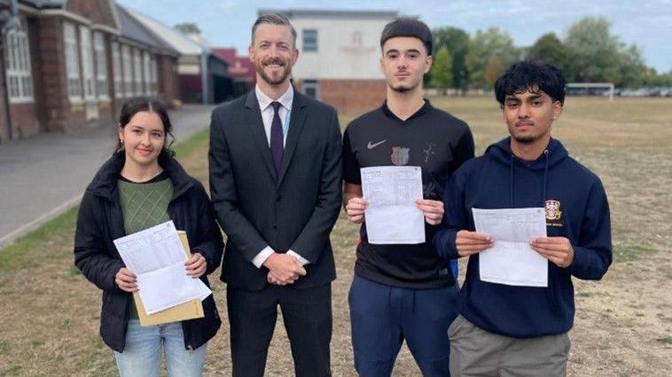 Headteacher Oliver King wears a grey two-piece suit, with a white shirt and dark purple tie. He has his hands clasped in front of him and greying dark hair swept into a side parting and a neatly shaved beard. He is standing with two male students to the right of the image, who are smiling and holding a piece of paper with their results on. A young female student stands to the left, she has brown curly hair tied in a ponytail and she is smiling holding her results.