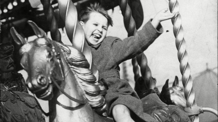 A black and white picture of a boy on a horse ride waving as it goes round. He has his hand out and his tongue. His hair is swept back.