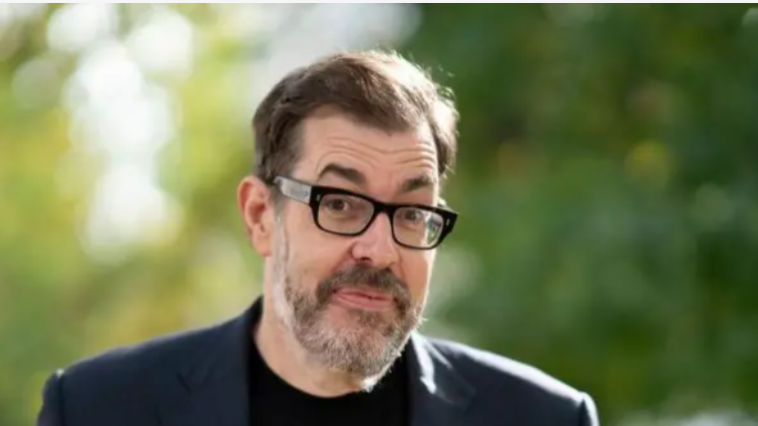 A picture of Richard Osman. He is wearing a navy suit with a black t-shirt and rectangular glasses with thick, black rims. He has dark hair and a grey beard. The background is out of focus trees.